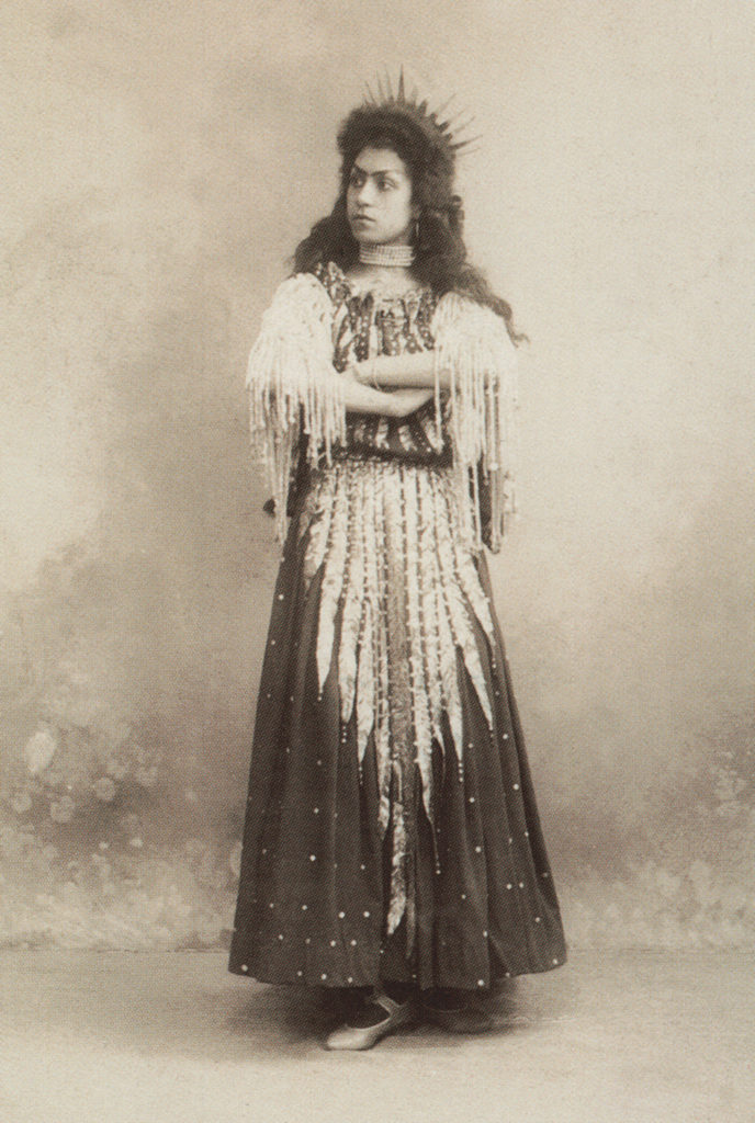Skorsiuk stands with her arms folded, looking over her right shoulder, in costume for the role of Morena in the Petipa ballet "Mlada." Her costume is a long, ornate dress with a tall crown. The photograph is sepia-toned.