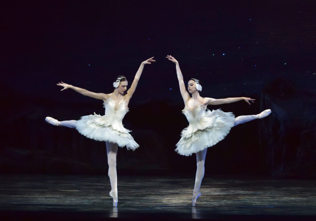April Giangeruso and Paulina Waski perform on a darkly lit stage in white swan tutus and feather headpieces with pink tights and pointe shoes. Their dark hair is slicked into low buns. They split center and arabesque en pointe toward mid-stage, into each other, heads inclined down with emotion. Their inside arms, in high arabesque line, create an archway as their outside arms follow the lines of their arabesque legs.