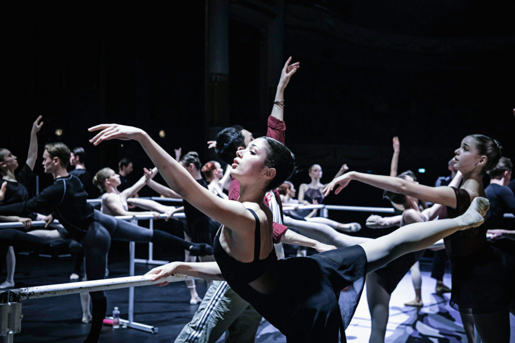 A female dancer is shown practicing second arabesque at a portable barre, lifting her right leg up. She is surrounded by other dancers doing the same step at multiple portable ballet barres lined up onstage. She wears a black leotard, black skirt, pink tights and ballet slippers.