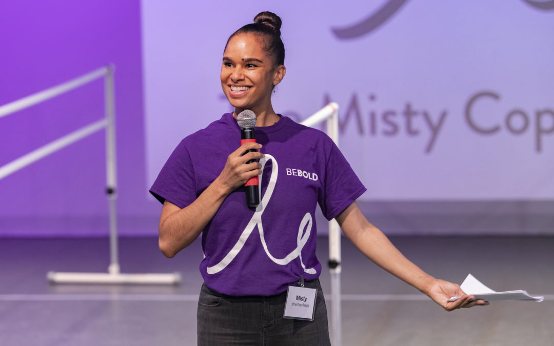 Misty Copeland speaks into a microphone during a demonstration. She wears a purple T-shirt and black jeans and holds a piece of paper in herleft hand..