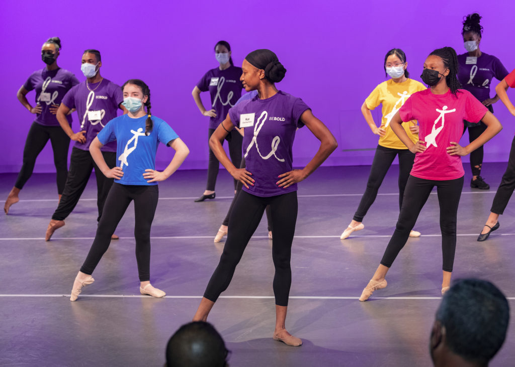 A ballet teacher stands at the front of a stage demonstrating tendu à la seconde with her hands on her hips. She turns to look at a large class of kids doing the same step behind her. They all wear brightly colored T-shirts and black leggings and ballet slippers.