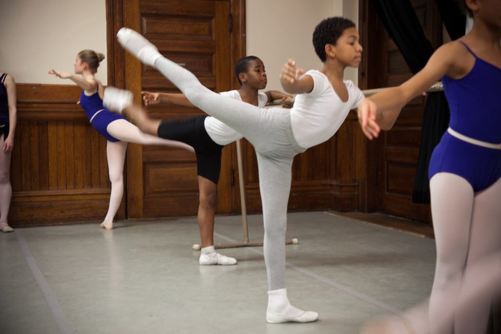 Victor Abreu stands at a ballet barre among other young ballet students and practices a grand battement derriere with his right leg high behind him. He holds his right arm in second position. He wears gray tights and a hite shirt, socks and ballet slippers.