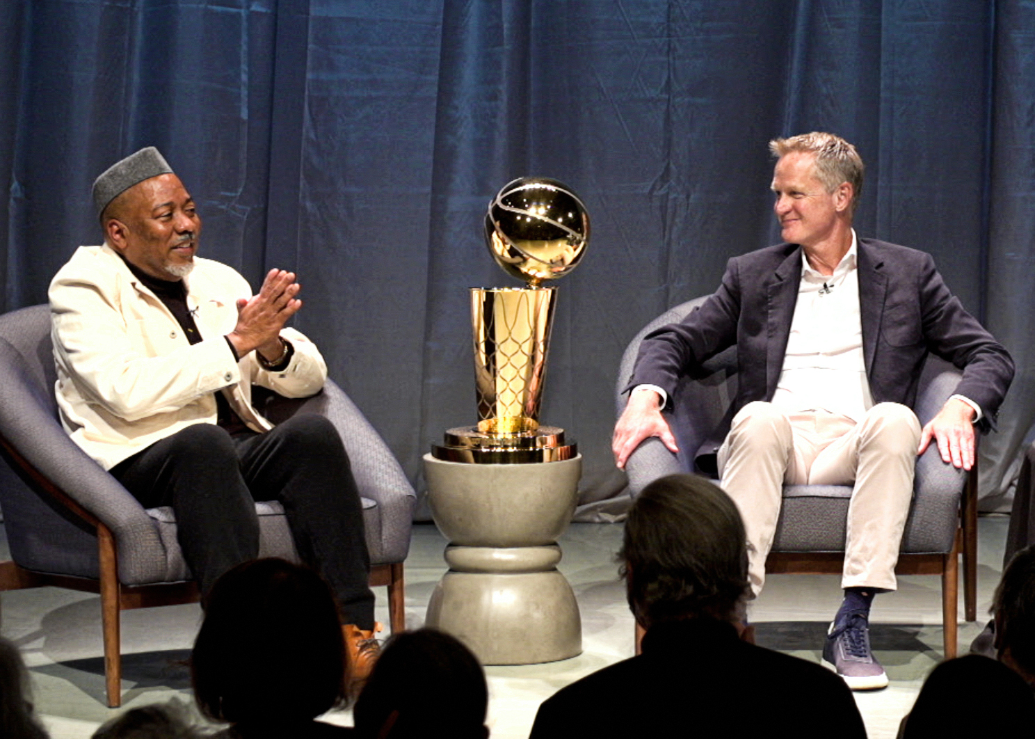 Alonzo King and Steve Kerr sit in gray-blue armchairs onstage during a public interview. Between them on a small table is a large gold NBA Championship trophy. King wears a short, light-colored jacket and black pants and a gray hat, and clasps his hands together in front of his chest. Kerr, on the right, wears a dark sport coat, white button-down shirt, khaki pants and blue tennis shoes, and looks toward King with a smile.