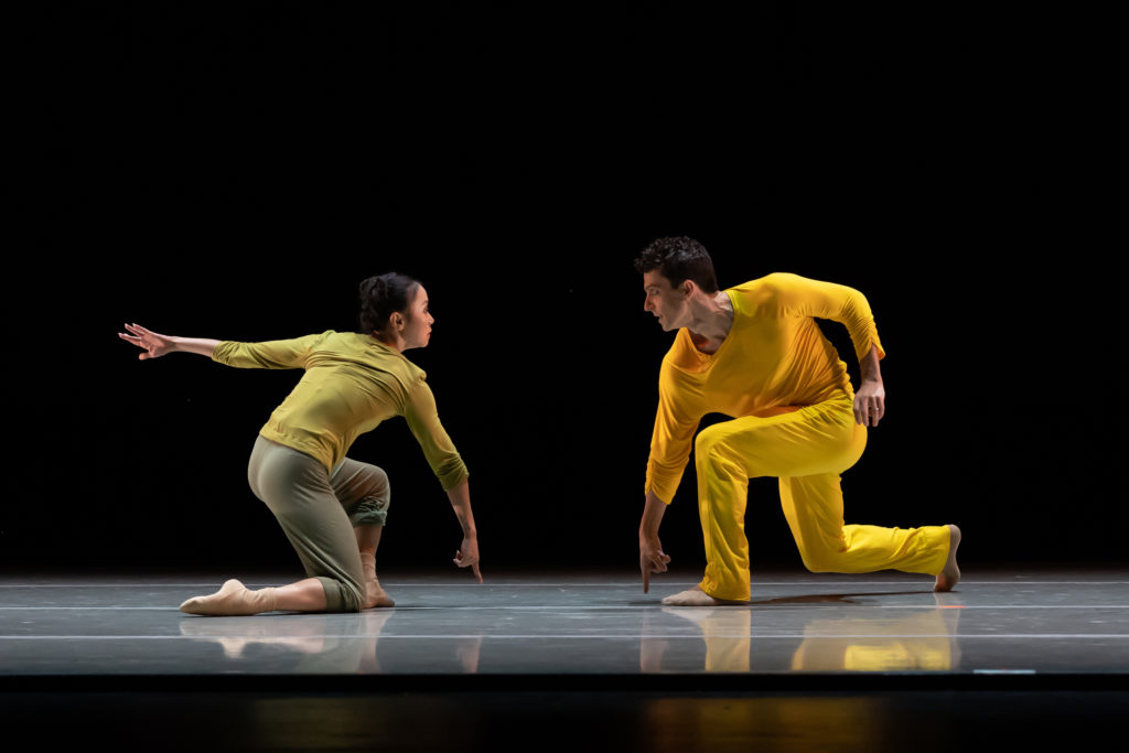 Cincinnati Ballet dancers Minori Sakita and Simon Plant perform onstage in front of a black backdrop. The dancers face into each other, kneel onto their right knee and point their right pointer finger down towards the ground. Minori Sakita, on the left, wears a green shirt and knee-length pants and think socks. Simon Plant wears a bright yellow pants and long-sleeved shirt and socks.