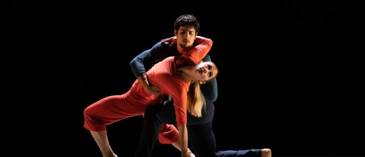 Melissa Gelfin De-Poli and Rafael Quenedit perform onstage in front of a black backdrop. Quenedit kneels down onto his right knee and holds Gelfin De-Poli around the waist as she wraps her arm around his left shoulder and leans back over his left thigh. They wear long-sleeved shirts and pants; Gelfin De-Poli's are orange and Quenedit's are dark blue.