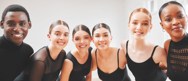 A group of six ballet dancers, five women and one man, stand huddled together and smile towards the camera. The women wear black leotards and pink tights and the man wears a black T-shirt and black tights.