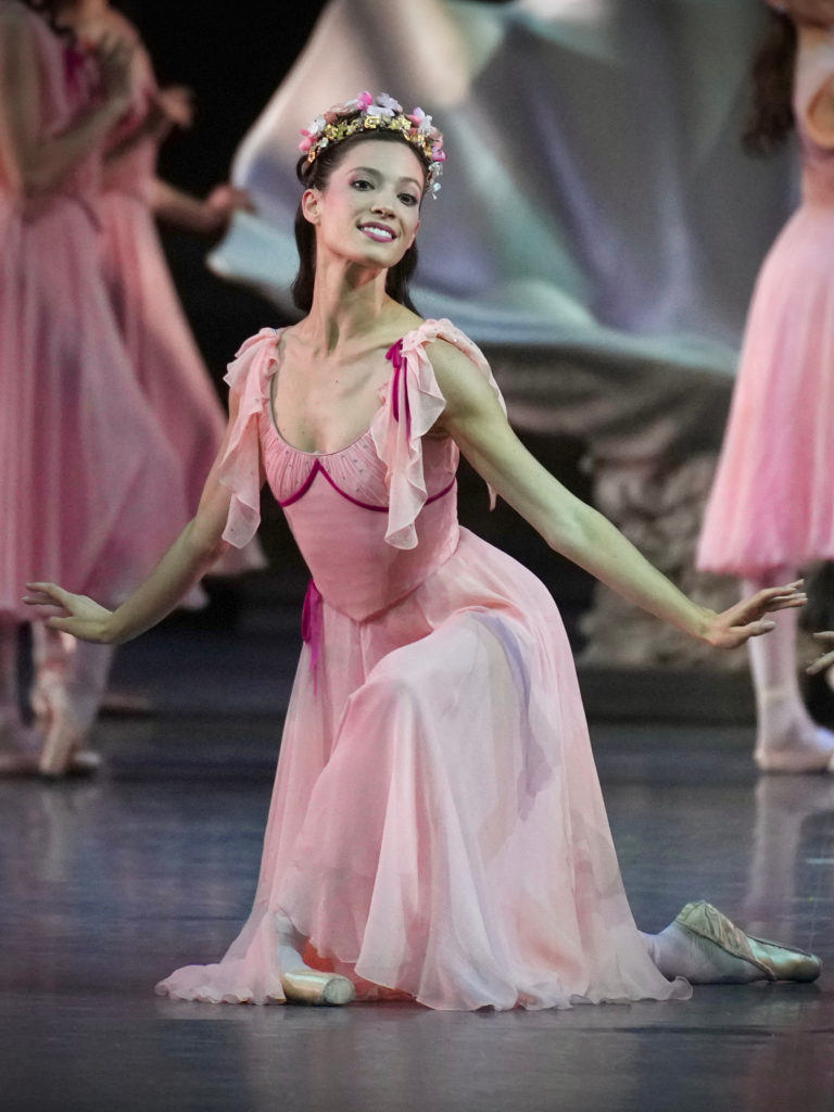 Lillian DiPiazza kneels on her right knee during a performance onstage. She wears a pink, billowy dress, pink tights, pointe shoes and a wreath of flowers on her head. She wears her dark hair long and smiles out toward the audience.