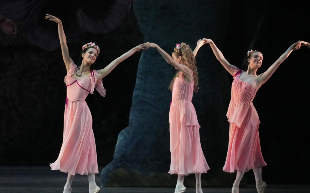 Lillian DiPiazza poses with two other dancers; they all lift their arms and hold hands, wearing calf-length pink dresses. The women wear their hair half up, half down and have a wreath of pink flowers in their hair.