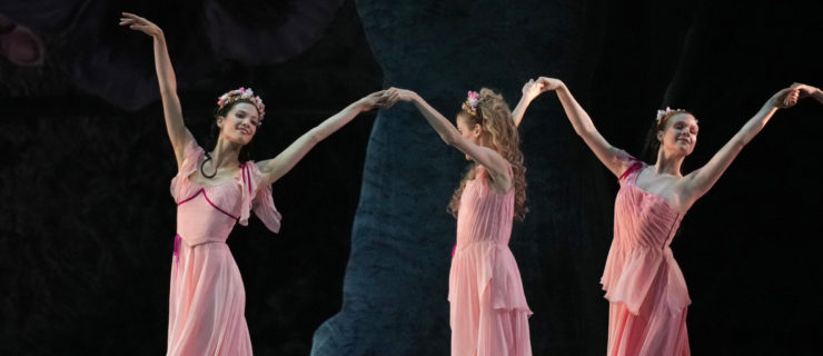 Lillian DiPiazza poses with two other dancers; they all lift their arms and hold hands, wearing calf-length pink dresses. The women wear their hair half up, half down and have a wreath of pink flowers in their hair.
