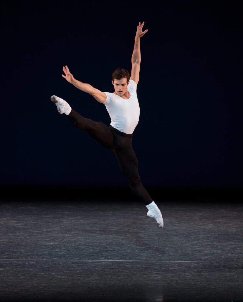 Nayon Iovino leaps onstage in an a la seconde sissone, his arms extended on the high diagonals. He wears a fitted white t-shirt, white socks and ballet flats, and black tights.