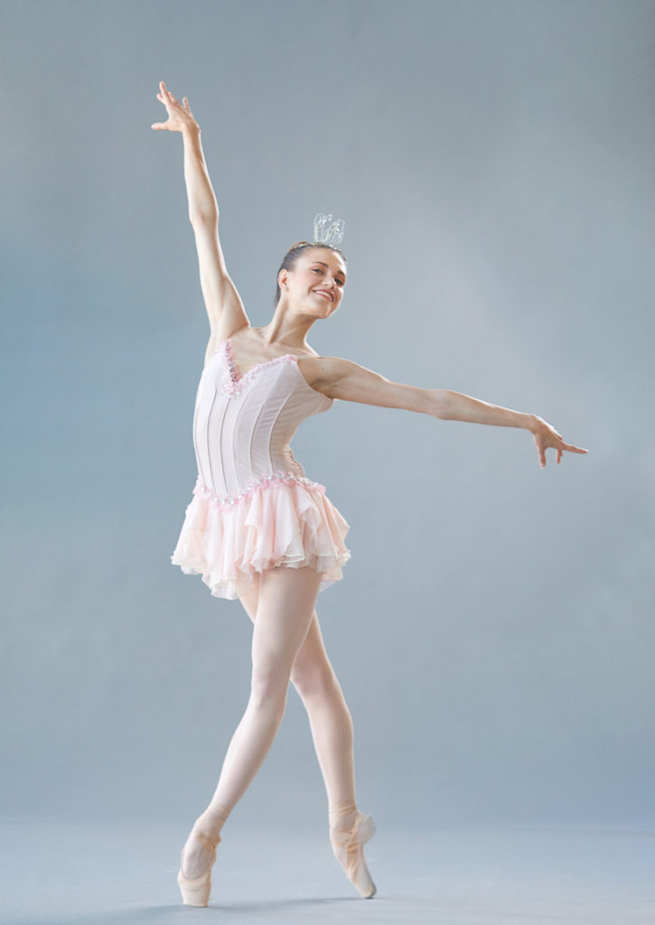Yulia Moskalenko, in a Dew Drop costume from The Nutcracker, stands in a wide fourth position croisé on pointe with her left foot in front. She holds her right arm up high and her left arm out to the side, and smiles widely as she looks out over her left shoulder.