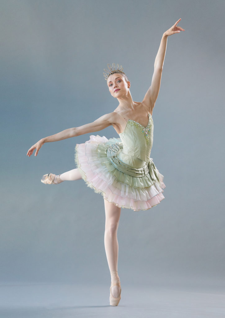 Yulia MOskalenko poses in a low fourth arabesque on pointe with her left leg back. She looks out towards the camera confidently and wears a short mint-green tutu with pink tulle trim and a delicate gold tiara.