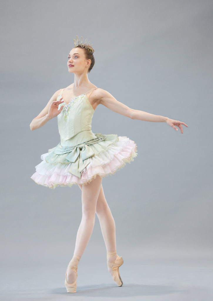Yulia Moskalenko wears a short mint-green tutu with pink tulle trim, a gold tiara, pink tights and pointe shoes. She poses in fourth position croisé on pointe with her left leg in front, her left arm out to the side, and her right hand pulled in towards her chest. She looks up towards the corner with a small, closed-mouth smile.