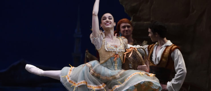 Betsy McBride performs a grand jeté onstage during a performance of Don Quixote. She wears a blue peasant dress with a peach bodice and peach trimmings, and holds a fan in her raised right hand and a basket with her left.