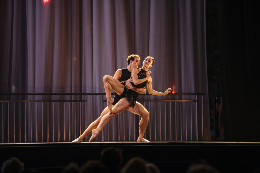 Betsy McBride and Jacob Clerico dance together during an evening performance on an outdoor stage. Clerico holds McBride around the waist and leans her to his left; she is in a passé position on pointe with her arms off to her left side and bent at the elbows and her face turned up. They both wear shot black costumes.