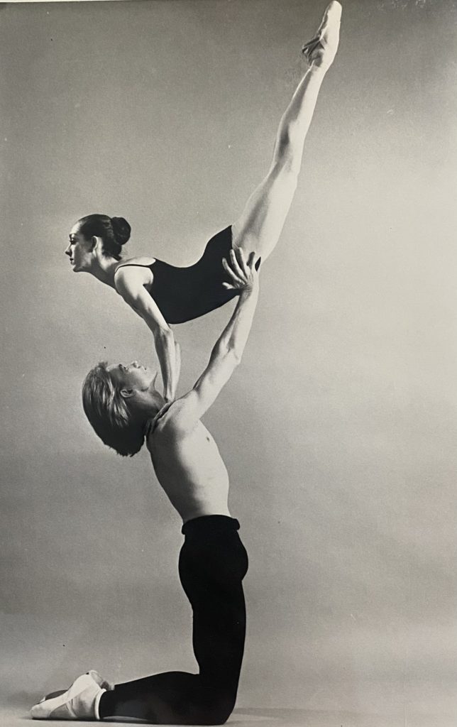 In this black and white photo, a male dancer in black tights, white socks and ballet slippers, kneels on both knees and lifts a ballerina Clara Cravey Stanley above his head, his hands on her hips as she points her legs up towards the sky. She presses her hands down on his shoulders and arches her back. She wears a black leotard, pink tights and pointe shoes.