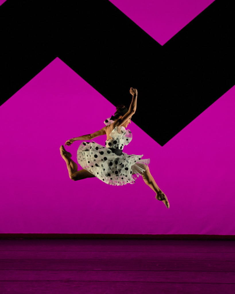 Betsy McBride jumps up with both knees bent and lifts her right arm up over her head. Behind her is a birhgtly lit purple backdrop with a black zig-zag design. She wears a white dress with black polka dots and tan ballet slippers.