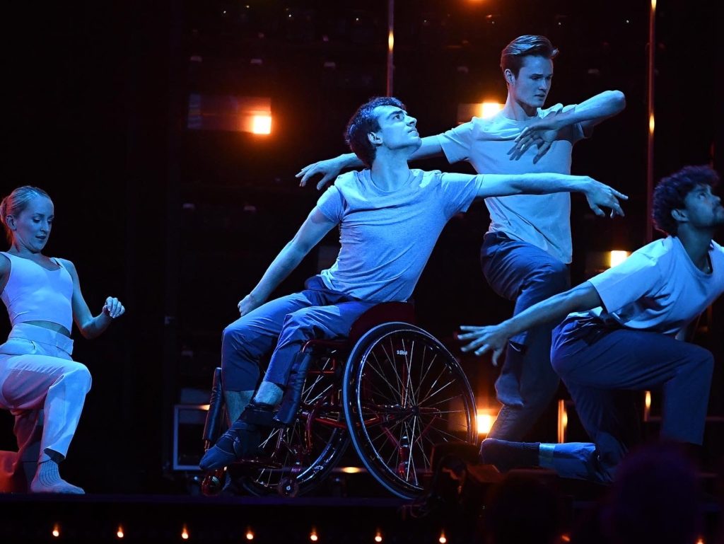 Joe Powell-Main reaches to the left with his upper body onstage during a performance, surrounded by stage lights and other dancers. He sits in a wheelchari and wears a light T-shirt and blue pants, black socks and black ballet slippers.