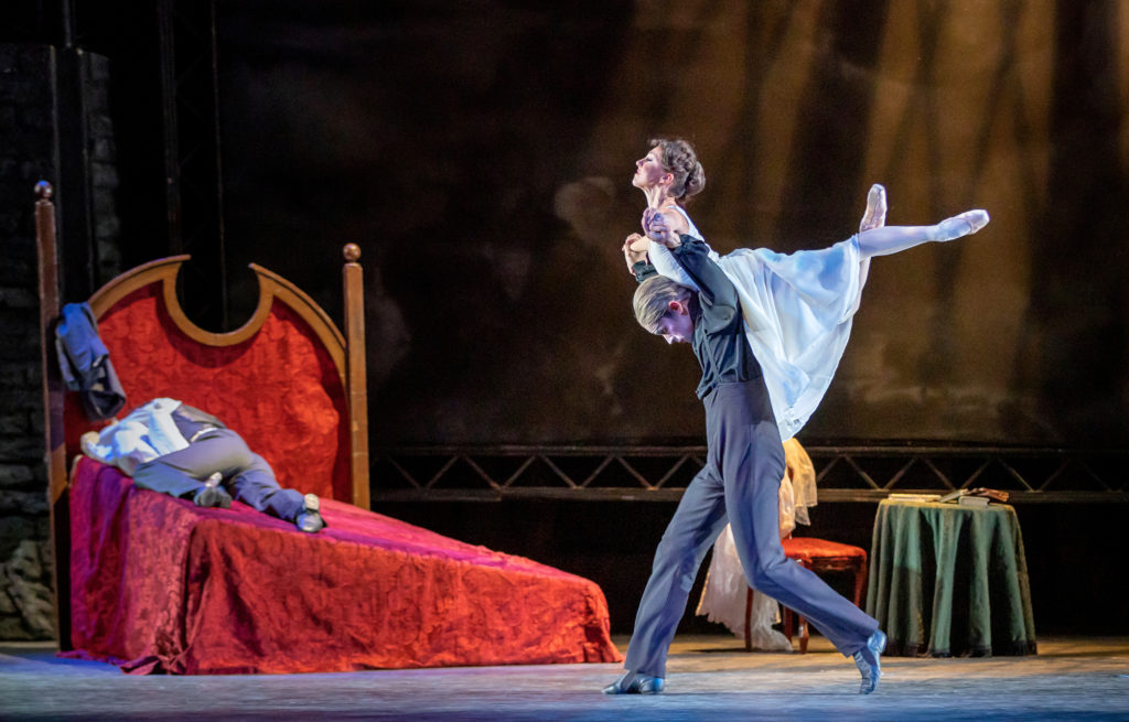 In a lavish bedroom scene onstage, a male dancer in a gray suit lies face down on a large bed with lush red sheets. In front of the bed, another male dancer dressed as Dracula walks forward as he lifts a female dancer in a white dress on his back. With her ankles crossed and arms supported to the side by Dracula's arms, the female dancer looks up passionately, as if flying.