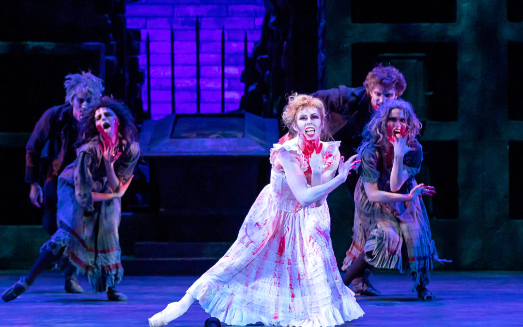 Jennifer Grace, in a long white dress stained with fake blood, dances onstage in front of a dark gated garden setting. She has darkened eye makeup, messy hair and fake bloodstains on her face and hands as she sneers at the audience, mouth wide open as if to lick the blood off of her hands. Two pairs of dancers, one male and one female per pair, dressed in darker colors mimic her movement and are also covered in fake bloodstains.