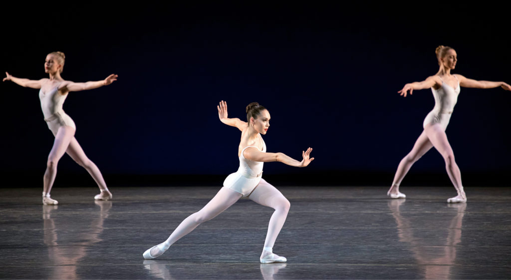 Mira Nadon dances a neoclassical ballet onstage in pink tights and pointe shoes and a white camisole leotard. She lunges and flexes her hands with her arms extended, her dark hair in a slick bun.
