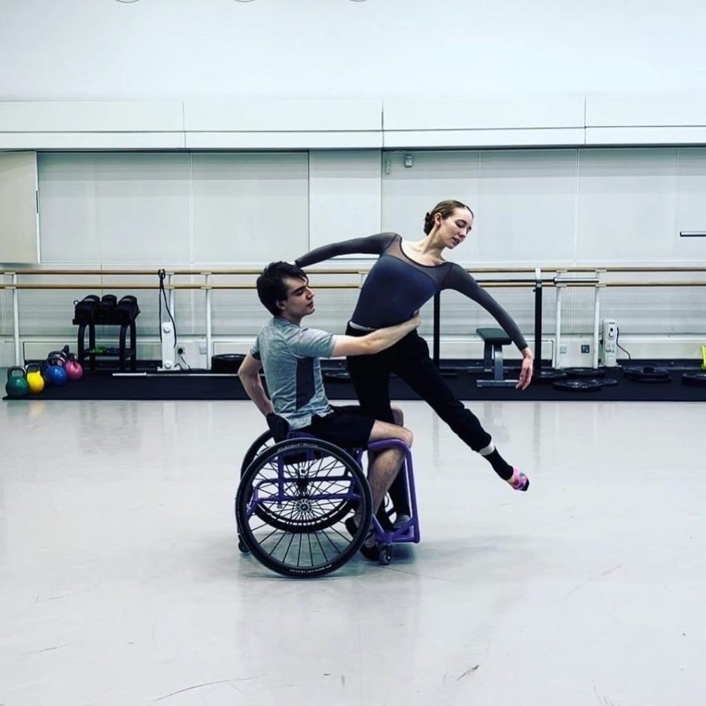 In a bright dance studio, Joe Powell-Main sits in a wheelchair and holds Isabel Lubach by the waist with his right arm as she balances on the chair's foot peddles on her right leg. She leans her upper body to the left and extends her turned in left leg out about 45 degrees. Lubach wears a blue long-sleeved leotard and black swetpants and Powell-Main wears a gray T-shirt and black shorts.