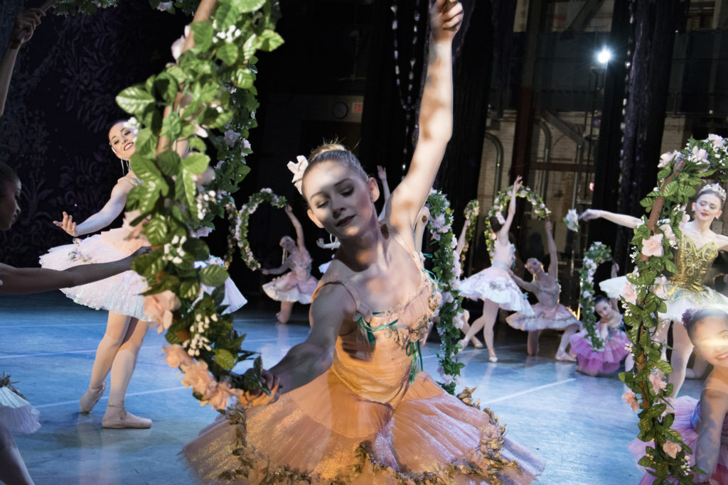 Jacqueline Callahan dances in the corps de ballet during a performance onstage. She wears a peach tutu and holds a large, leafy garland over her head as she arches her upper body over to her right. Other dancers make similar poses behind her, forming a circle, as soloists dance in the middle.