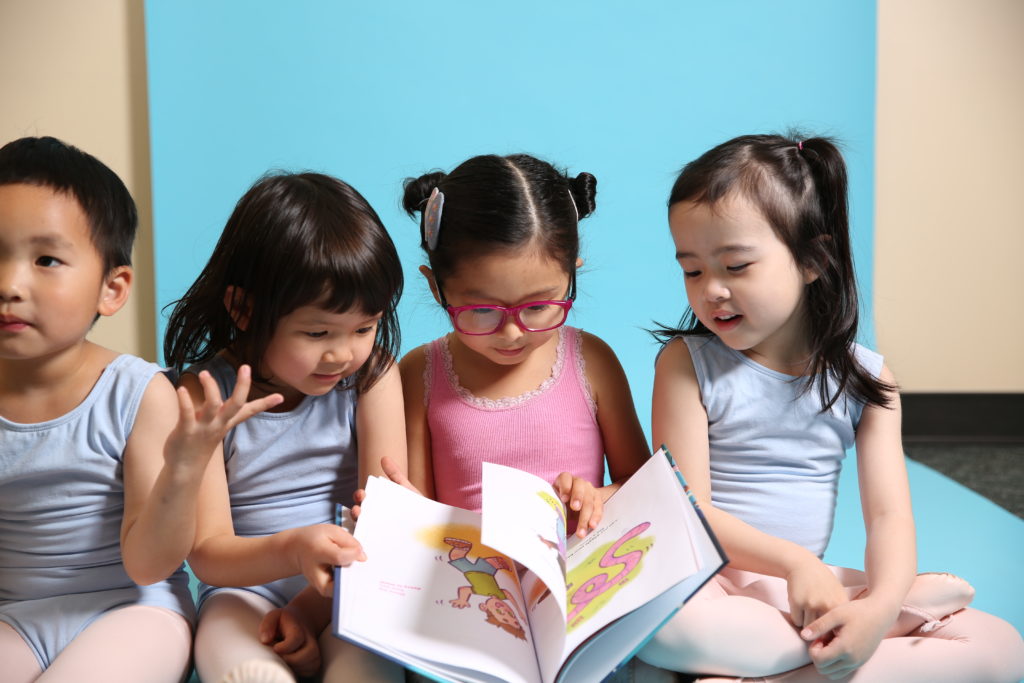 Four small children in leotards, tights and ballet shoes sit next to each other on the floor and page through a children's book.