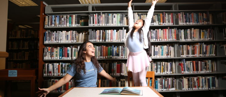 A college-age woman and girl ballet student are in a library at a bale in front of shelves of books. The little girl, dressed in a blue leotard, pink tights, pink skirt and white cardigan sweater stands on her chair and reaches her arms up above her head while lookig up toward the ceiling with a huge smile. The college-age dancer sits next to her and opens her arms wide to the side and makes jazz hands. She wears a blue T-shirt and smiles toward the little girl.