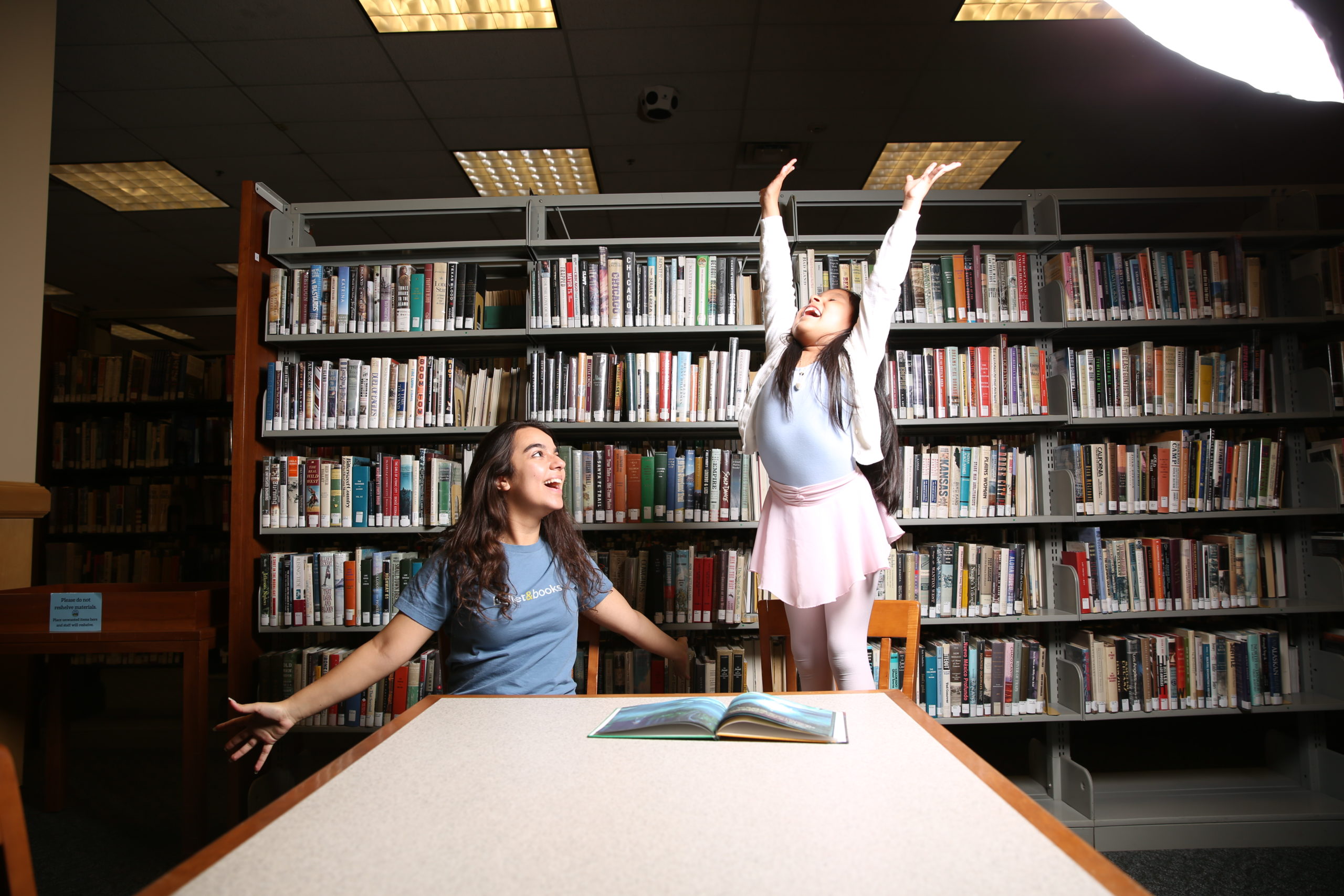 A college-age woman and girl ballet student are in a library at a bale in front of shelves of books. The little girl, dressed in a blue leotard, pink tights, pink skirt and white cardigan sweater stands on her chair and reaches her arms up above her head while lookig up toward the ceiling with a huge smile. The college-age dancer sits next to her and opens her arms wide to the side and makes jazz hands. She wears a blue T-shirt and smiles toward the little girl.