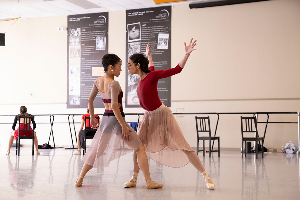 Maine Kawashima and Jaimi Cullen rehearse a dramatic scene in a large ballet studio. Kawashima, in a red leotard with long mesh nude-colored sleeves, black shorts, a long light pink dance skirt and pointe shoes, grounds her feet and clenches her fists, staring intensely at Cullen. Cullen, in a long-sleeved red leotard, nude tights, a light pink dance skirt and pointe shoes, leans toward her with emotion, her arms raised behind her with her hands flexed, as if about to grab or strike Kawashima. Dancers sit, leaning over, on black chairs behind them.