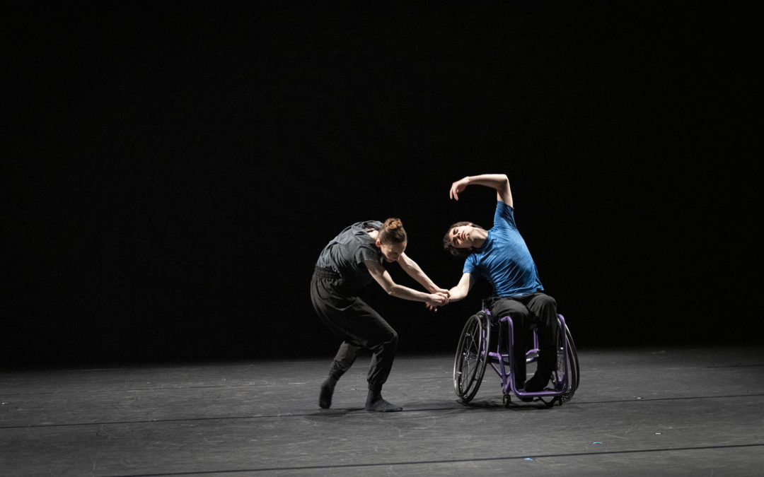 Joe Powell Main sits in a purple wheelchair and performs a cambré side to the right, with his left arm over his head. He wears a blue T-shirt and black sweatpants. Kristen McNally, in gray, loose-fitting dance clothing and dark socks, holds his forearm with both hands and bends her upper body forward as she takes a step toward Powell-Main. They dance onstage in front of a black backdrop.