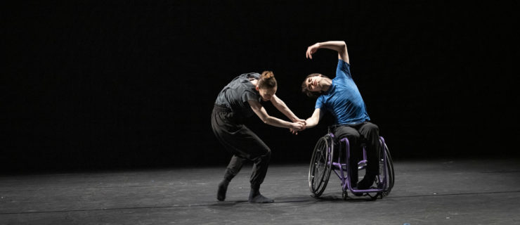 Joe Powell Main sits in a purple wheelchair and performs a cambré side to the right, with his left arm over his head. He wears a blue T-shirt and black sweatpants. Kristen McNally, in gray, loose-fitting dance clothing and dark socks, holds his forearm with both hands and bends her upper body forward as she takes a step toward Powell-Main. They dance onstage in front of a black backdrop.