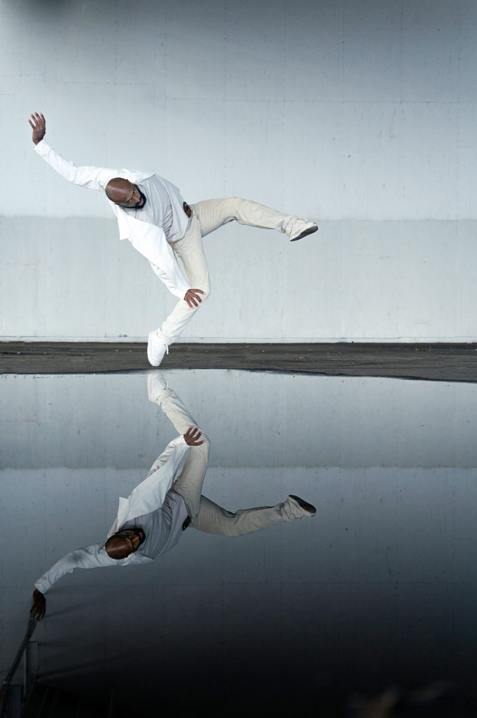 Babatunji Johnson wears white pants, sneakers and a white jacket. He leans back and balances on his right tow as he lifts his left leg up in attitude devant, spiraling his upper body. He poses in front of a white backdrop and looks into a pool of water, which reflects his image.