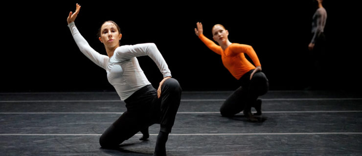 Brooke Corrales and Amelia Shultz crouch low to the ground, kneeling onto their right knee and pushing forward on their left foot while leaning back and lifting their right arm. They both wear black pants, black socks and long-sleeved tops. Ben Simeons walks across the stage behind them.