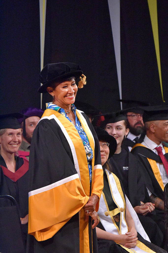 Julie Felix is standing onstage wearing a black cap with a yellow tassel and a black and yellow gown. She is surrounded by others from Falmouth University, also wearing caps and gowns.