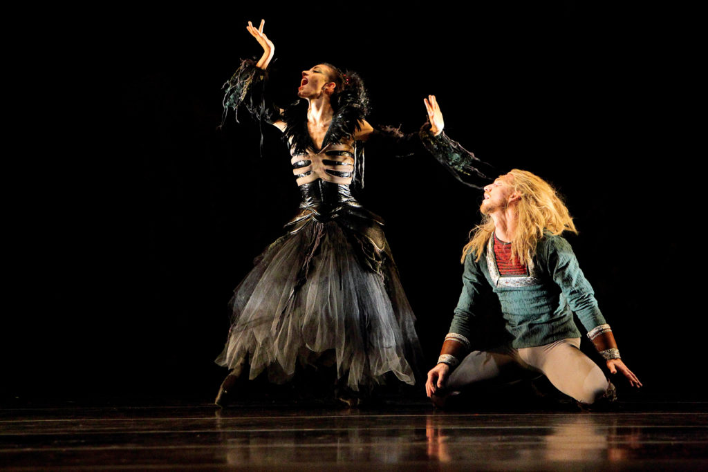 Onstage, a female dancer in a long black gothic dress with white skeletal decoration across her ribcage makes a dramatic gesture with her hands, mouth open, wearing a wild black wig. A male dancer, on his knees behind her, wears a blue tunic with a red undershirt, gray tights and a wild blonde wig. He looks up at her intensely.