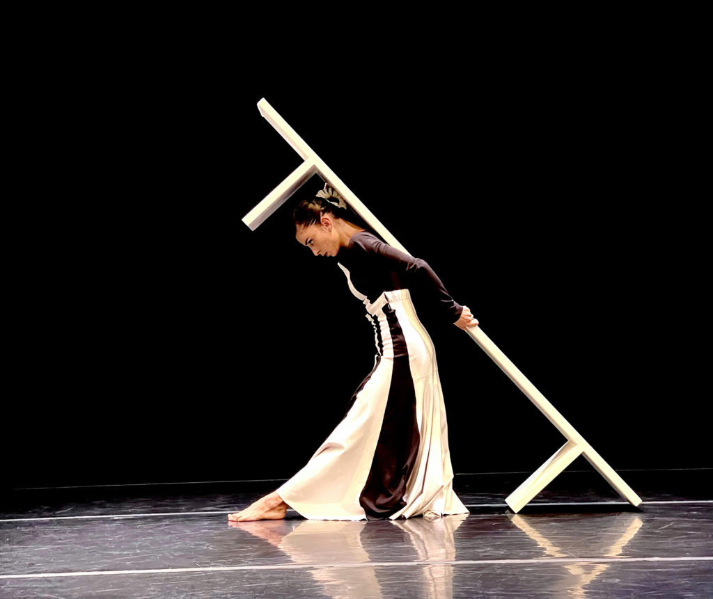 During a performance onstage, Dores André leans her body forward and holds a long white bench against her back. She wears a long dress with white and black panels.