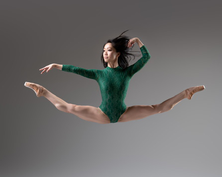 Erina Tanaka jumps into a second position split in the air. She extends her right arm out to the side and touches her hair wit the back of her left hand. She wears a green turtleneck-style leotard and tan pointe shoes.