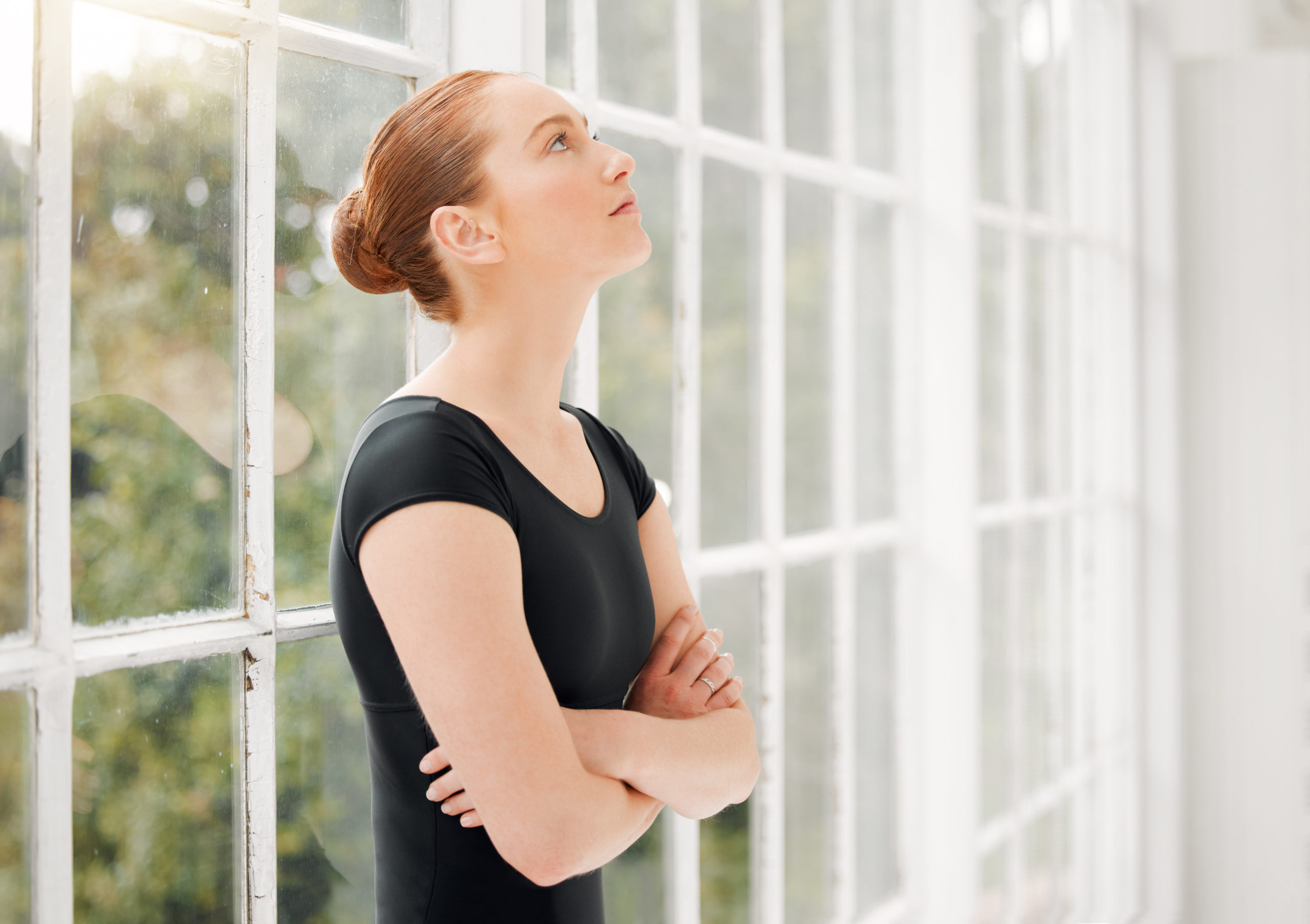 A female ballet dancer stands alone in front of a large windo, looking nervous and stressed
