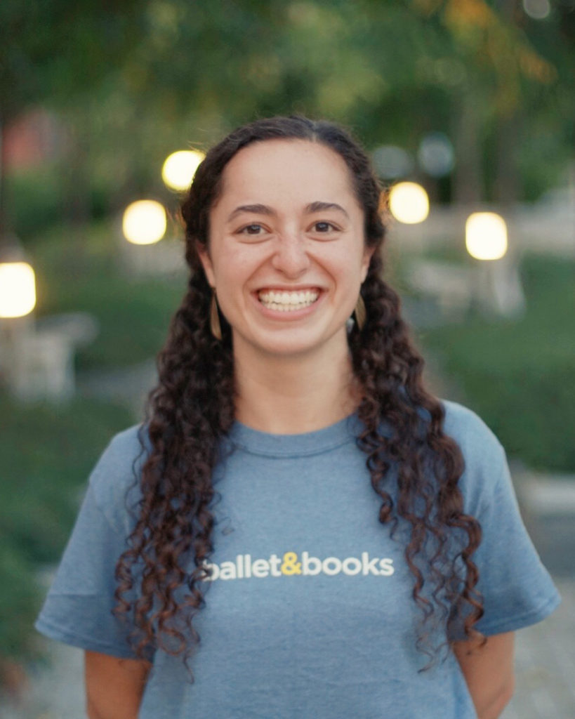 Talia Bailes wears a blue T-shirt with the words "ballet & books" written in small lettering across the front. She wears her curly hair down and smiles widely for the camera.