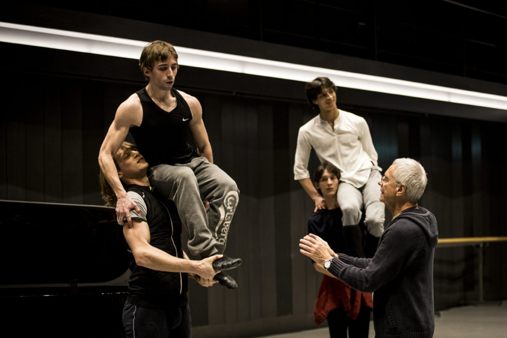 John Neumeier, wearing a gray hooded sweatshirt, works with two pairs of male dancers during a rehearsal as they practice a shoulder lift.