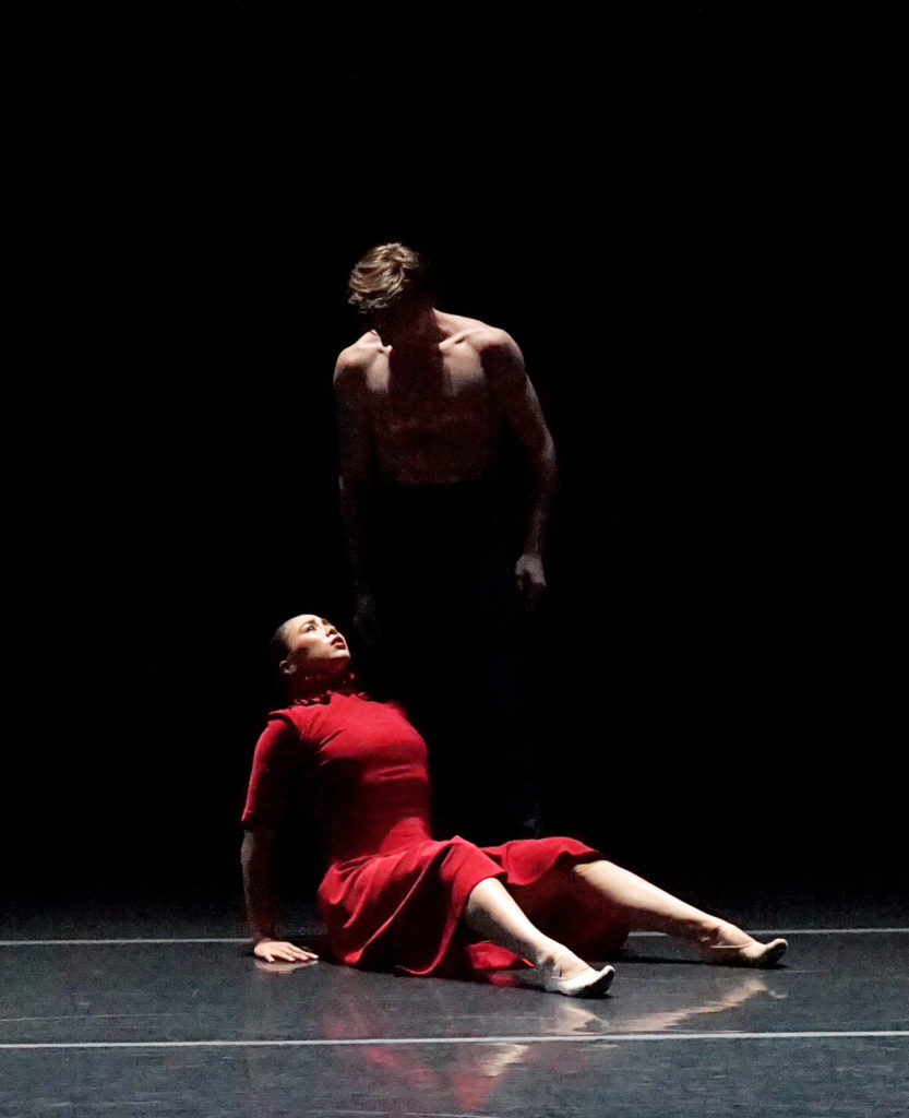 Katerina Beckman, wearing a bright red dress, sits on the floor onstage, leaning on her hands and with her knees bent, and looks up towards Ben Simoens, who stands directly behind her in the shadows.