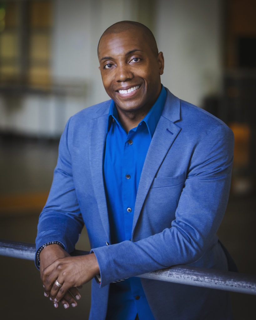 Kiyon Ross, dressed in a cobalt blue suit and button-down shirt, is shown waist-up as he leans over a ballet barre, smiling at the camera.