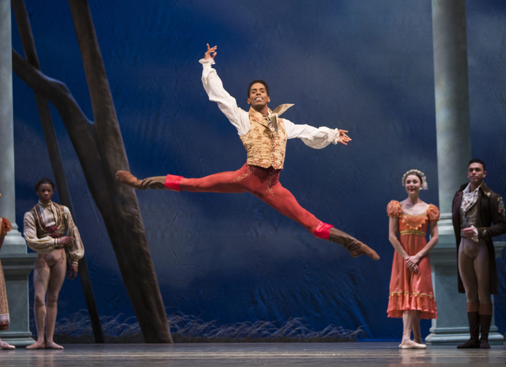 Jonathan Batista performs a saut de chat to his left during a performance while other dancers stand behind him and watch. He wears a white long-sleeved shirt and tan vest, coral colored tights and brown ballet boots.