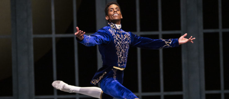 Jonathan Batista smiles broadly and does a grand jeté en evant with his back leg in attitude and his arms in second during a performance of Swan Lake. He wears a blue velvet prince costume with gold trim with white tights and white ballet slippers.