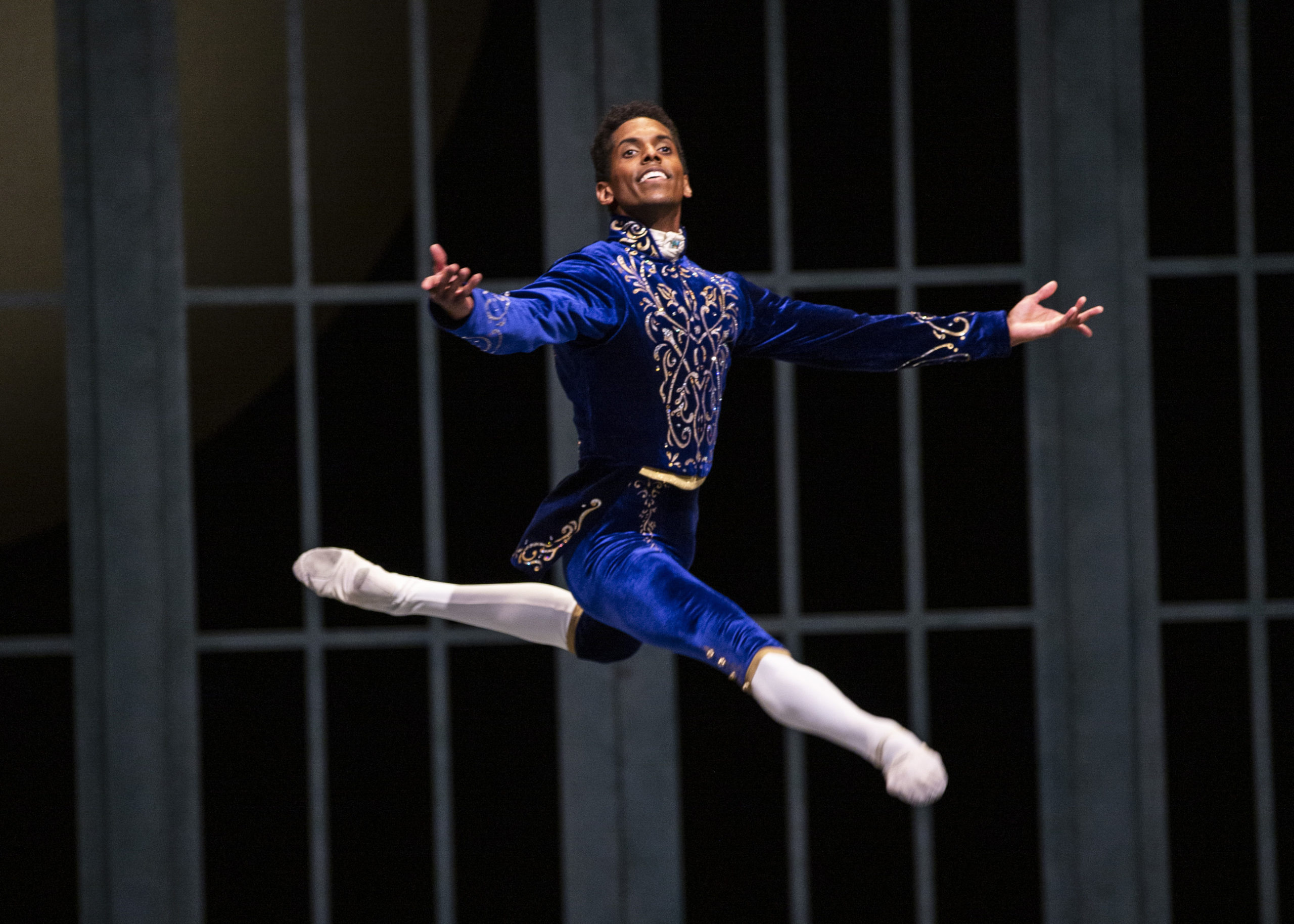 Jonathan Batista smiles broadly and does a grand jeté en evant with his back leg in attitude and his arms in second during a performance of Swan Lake. He wears a blue velvet prince costume with gold trim with white tights and white ballet slippers.