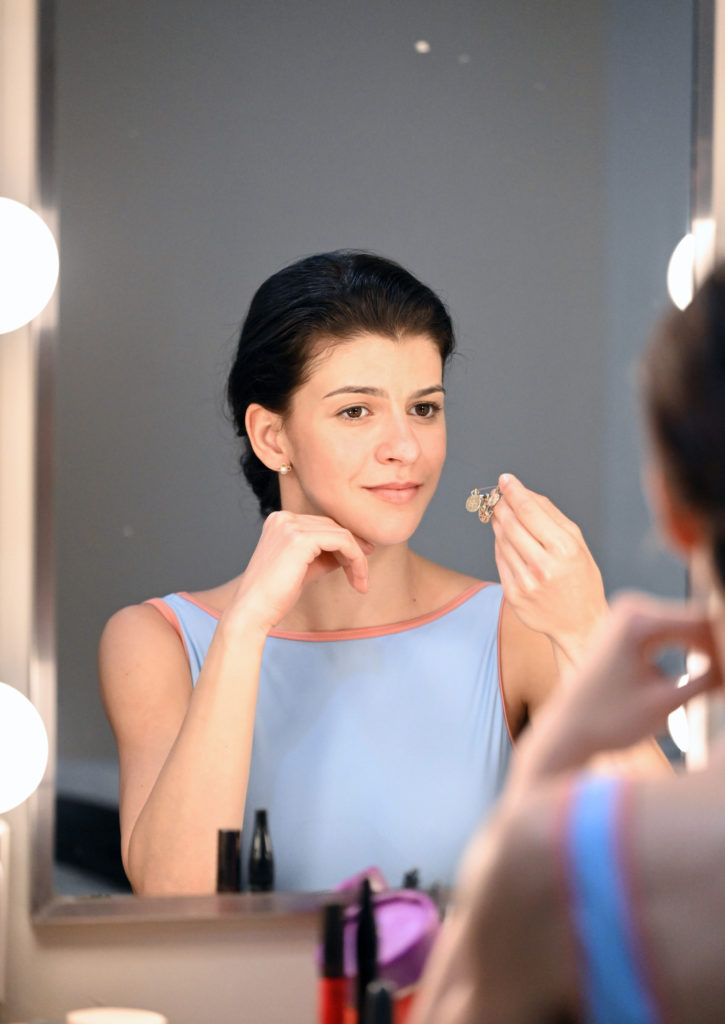Magri, wearing a light blue leotard, sits in front of a makeup mirror and holds her golden charm pin.