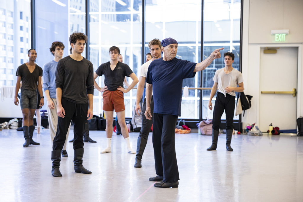 A group of 8 male dancers stand in a large dance studio, led by an older male repetiteur. They wear t-shirts, tights, workout pants and other athletic clothing, as well as ballet shoes. They all look toward the right side of the image and the repetiteur points in that direction.