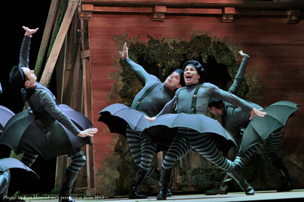 Abreu is onstage in a performance of "Platée," surrounded by castmates. He is wearing a green long-sleeved shirt, a green beret, black and green striped pants, and black boots. He is also wearing an umbrella-shaped tutu held by suspenders.